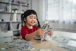 Asian baby girl wearing a red t-shirt holding dollar bill on wood table desk in living room at home. Saving investment wealth, growth stock, invest interest, fund, saving money for the future concept. photo