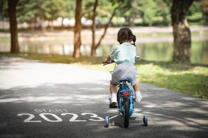 feliz año nuevo 2023,2023 simboliza el comienzo del nuevo año. la carta comienza el nuevo año 2023 en la bicicleta de ciclismo de la niña en la carretera en el jardín del parque natural. meta del éxito. fondo de pantalla del número 2023. foto