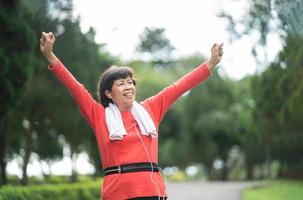 Athletic asian senior woman 60s smiling, hands up and happy cheerful with success and jogging. Senior asian woman running at park. Female runner listening to music with earphones while jogging. photo