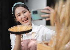 Young woman wears apron taking selfie photo with homemade pie in kitchen. Portrait of beautiful Asian female baking dessert and having fun taking photo by smartphone for online social. Home cookery.