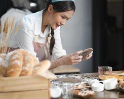 Young woman wears apron taking selfie photo with homemade pie in kitchen. Portrait of beautiful Asian female baking dessert and having fun taking photo by smartphone for online social. Home cookery.