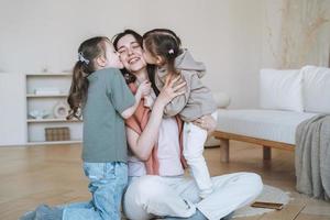 joven familia feliz con una madre madre con dos hijos niñas besando a mamá en la sala de estar en casa foto