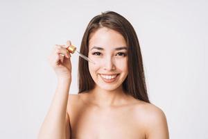 retrato de belleza de una mujer asiática feliz y sonriente con el pelo largo y oscuro con pipeta con aceite orgánico en la cara y las manos limpias y frescas de la piel sobre fondo blanco aislado foto