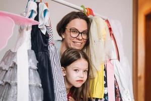 linda niñita con el pelo largo y oscuro vestido de rojo con madre joven con gafas entre sus hermosos vestidos en el armario de la habitación de los niños en casa foto