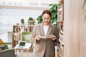 Adult smiling brunette business woman forty years with long hair in stylish beige suit and jeans at public place, green open space office, coworking. Friendly teacher or mentor with book in library photo