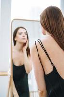 mujer joven con el pelo largo y oscuro en un elegante vestido negro de noche con la espalda abierta cerca del espejo en el interior blanco, gente por detrás foto