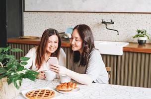 amigas morenas adultas sonrientes desayunando y usando el teléfono móvil en la cocina de casa foto