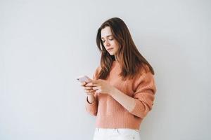 Portrait of beautiful smiling woman student with dark long hair in casual knitted sweater using mobile phone in hand on grey background photo