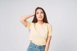 joven mujer asiática infeliz con cabello largo en camisa amarilla y jeans sosteniendo sus manos en la cabeza sobre fondo gris foto