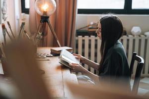 joven sonriente mujer adolescente estudiante con cabello largo oscuro en un libro de lectura informal en casa foto