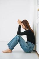 joven mujer infeliz adolescente con el pelo largo en jeans sentada en el suelo en posición cerrada junto a la puerta de casa, emoción negativa