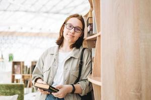 Young brunette teenager girl student in glasses with mobile phone at modern library, public place photo