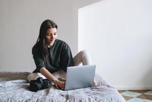 joven fotógrafa con cámara digital independiente vestida de forma casual usando una laptop sentada en la cama en casa foto