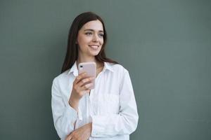 Portrait of beautiful smiling woman student with dark long hair in white shirt using mobile phone in hand on grey background isolated, generation Z photo