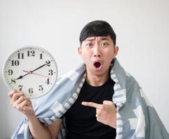 Young Asian man shocked and point finger at clock in his hand with the blanket cover body and looking at camera on white isolated work late concept photo