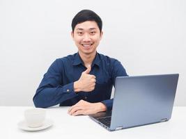 el hombre alegre sonríe y levanta el pulgar con su computadora portátil sobre la mesa foto