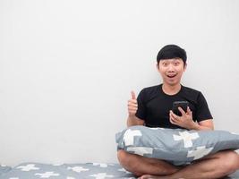 Asian man holding mobile phone and thumb up on the bed copy space photo