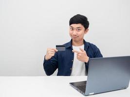 hombre feliz emoción sentarse con la computadora portátil en la mesa señalar con el dedo la tarjeta de crédito en la mano foto