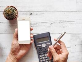 Hand holding cellphone white screen and using calculator with cactus and silver pen on white wood table nature shadow and sunlight top view and space business concept photo