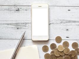 Smartphone white display silver pen on blank page notebook and gold coins group business concept,Business gadget for calculate on white wood table top view photo