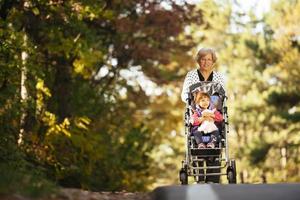 Happy senior lady pushing wheel chair and children. Grandmother and kids enjoying a walk in the park. Child supporting disabled grandparent. Family visit. Generations love and relationship photo