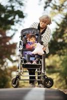 Happy senior lady pushing wheel chair and children. Grandmother and kids enjoying a walk in the park. Child supporting disabled grandparent. Family visit. Generations love and relationship photo