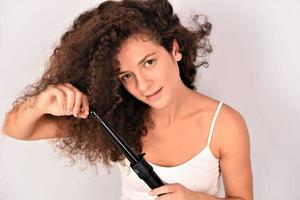 Pelo RIZADO. bella mujer sonriente con el pelo largo y ondulado planchándolo, usando un rizador. chica feliz con un hermoso cabello suave y saludable usando un rizador para rizos perfectos. peinado y peluqueria foto