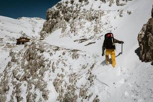 hombre solitario sube boca arriba a través de la nieve profunda hasta la cabaña de montaña en el borde de la montaña usando el piolet durante el soleado día de primavera en los alpes italianos en europa foto