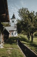 Old white church with tower on the edge of the old Slovenian village with the stream flowing around and shadows below the trees in garden during the hot summer sunny day in vertical orientation photo