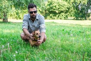 apuesto hombre europeo con estilo sentado en la hierba con su perro en el parque en un paseo. amistad entre humanos y mascotas.