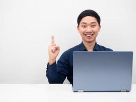 Un hombre guapo con una laptop en la mesa, una sonrisa feliz, tiene una idea, apunta con el dedo hacia arriba en el espacio de la copia, blanco aislado. foto