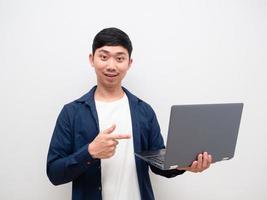 hombre asiático camisa azul alegre apuntar con el dedo a la computadora portátil en la mano y mirar la cámara sobre fondo blanco aislado foto