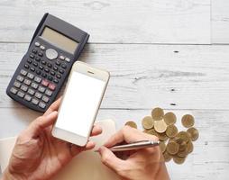 Hand with silver pen using calculator and holding smartphone white screen with gold coins group on white wood table top view and copy space nature shadow photo