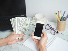 mano sosteniendo dinero en dólares y teléfono móvil con espacio de trabajo en el fondo de la mesa de madera blanca foto