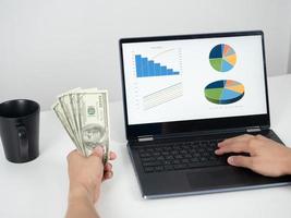 mano de hombre sosteniendo dinero en dólares y usando una computadora portátil en la mesa con una taza de café foto