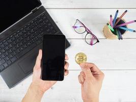 Hand holding mobilephone and gold bitcoin with laptop glasses and pen box background top view photo