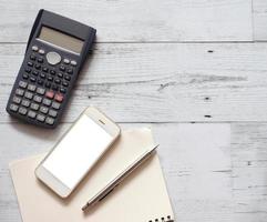 Smartphone white screen silver pen on blank page notebook with calculator on white wood table top view and copy space photo