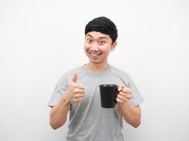 hombre asiático sonriendo y sosteniendo una taza de café y pulgar arriba blanco aislado foto