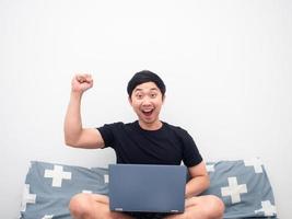 Man sitting one the bed with laptop feeling happy show fist up white background photo