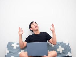 Man sitting on the bed with laptop feeling excited and looking up at copy space photo