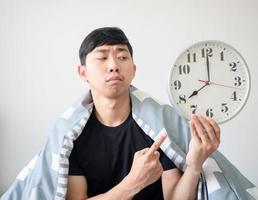Young man with blanket cover his body looking and point finger at clock in hand feel bored at face on white isolated