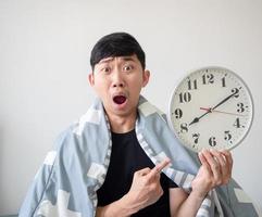 Asian man cover blanket point at clock in his hand with shocked face late concept white background photo