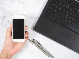 Top view workspace hand holding mobile phone with silver pen and laptop white marble desk photo