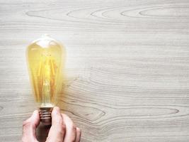 Hand holding yellow light bulb on wood texture table copy space top view,Inspiration economy concept photo