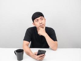 hombre sentado a la mesa con gesto de taza de café pensando y sosteniendo gesto de teléfono móvil pensando mirando hacia arriba foto