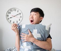 Asian man shocked face and looking at clock in hand and hugging pillow on white isolated wake up late concept photo