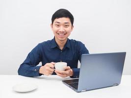 Asian man holding coffee cup with happy smile at his workplace laptop on the table photo