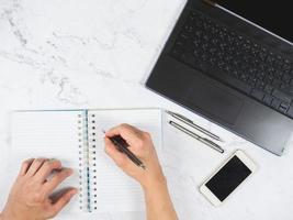 vista superior espacio de trabajo mesa de mármol escritura a mano en un cuaderno con bolígrafo y teléfono móvil y espacio para computadora portátil foto