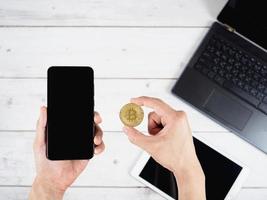 Hand holding gold bitcoin and mobilephone with laptop and tablet on wood table background photo