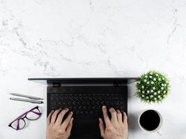 mano en el teclado portátil con gafas pluma plateada taza de café florero en mesa de mármol concepto de lugar de trabajo espacio de copia vista superior foto